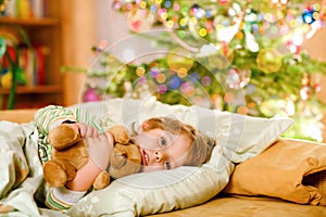 Little cute blond boy sleeping under Christmas tree and dreaming of Santa at home, indoors. Traditional Christian