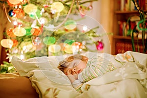Little cute blond boy sleeping under Christmas tree and dreaming of Santa at home, indoors. Traditional Christian