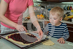 Little cute blond boy watches with curiosity as his mother spreads jam on dough for baking berry pie. Joint family cooking at home
