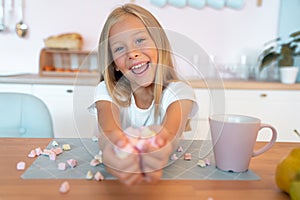 Little cute blond girl in the kitchen having her breakfast having fun with marshmallow and smiling. This is so tasty