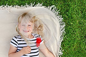 Little cute blond girl lying on blanket over green grass lawn and smiling. Adorable child having fun outdoors. Happy family, paren