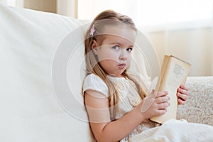 Little cute blond girl reading book siting on a sofa. Child reading, dreaming and imagination development