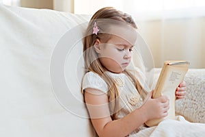 Little cute blond girl reading book siting on a sofa. Child reading, dreaming and imagination development