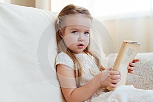 Little cute blond girl reading book siting on a sofa. Child reading, dreaming and imagination development