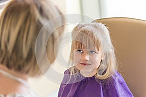 Little cute blond girl smiles and looks at hairdresser during haircut process.