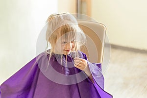 Little cute blond girl smiles and looks at hairdresser during haircut process. Girl holds a wet forelock hair before it will be al