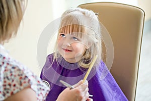 Little cute blond girl smiles and looks at hairdresser during haircut process. Hairdresser holds comb and scissors in a hand