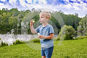 Little cute blond kid boy having fun play blowing soap bubbles at park near lake on bright warm summer spring day.happy healthy