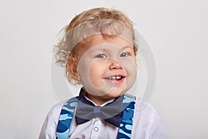 Little cute blonde boy wearing bow tie and blue camouflage suspenders, looking directly at camera with toothy smile, posing