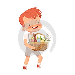 Little cute boy carries a basket with a chocolate hare, colored eggs and osier branches for the Easter holiday. Spring