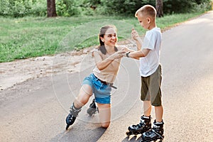 Little cute boy help mother to stand up from road after falling down in roller skates, smiling woman looking at her child, family