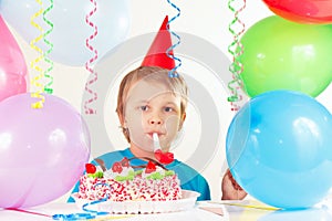 Little cute boy in holiday hat with birthday cake with whistle and festive balloons