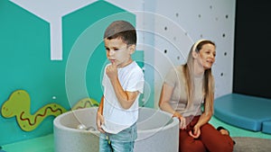 little cute boy in jeans and white t-shirt thinking in the playroom, teacher and balls in the background