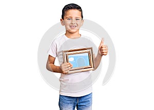 Little cute boy kid holding empty frame smiling happy and positive, thumb up doing excellent and approval sign