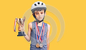Little cute boy kid wearing bike helmet and winner medals holding winner trophy thinking attitude and sober expression looking