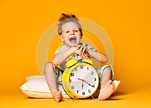 Boy in pajamas holding a toy dinosaur in his hands, sitting on a pillow with an alarm clock.  on a yellow background.