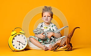 Little cute boy in pajamas holding a toy dinosaur in his hands, sitting on a pillow with an alarm clock. Isolated on a yellow