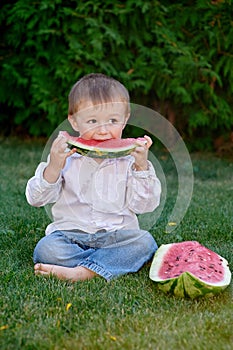 Little cute boy sitting on grass in park and eating a watermelon