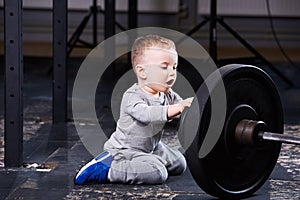 Little cute boy in sportwear sitting on the floor at the gym and looking on the barbell.