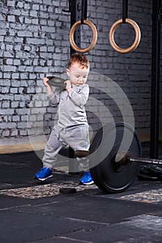 Little cute boy in sportwear standing with disk of the barbell at the gym and looking on the barbell.