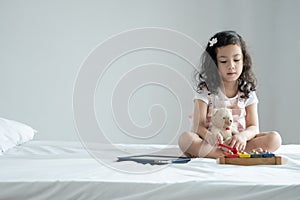 Little cute Caucasian Asian kid sitting on bed concentrating on playing small colorful xylophone with bear doll and tablet compute