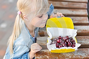 Little cute caucasian blond girl looking at lunchbox with fresh tasty sweet cherries on wooden table outdoors.Kid going to eat swe