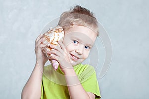 Little cute caucasian boy with blond hair and blue eyes listens to seashell, smiling.