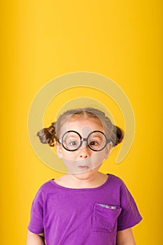 Little cute caucasian funny surprised kid child baby girl with glasses in purple T-shirt on yellow wall background