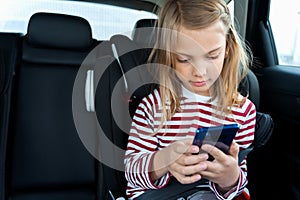 Little cute caucasian girl is driving in car. Kid writing sms, chatting, playing video games in mobile phone. Traveling on road in