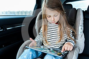 Little cute caucasian girl is driving in car. Online school learning,education.Playing video games in tablet. Traveling on road in