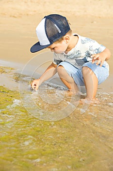 Little cute caucasian toddler boy with blond hair plays at the beach in sea waves on sunny summer day. Family vacation with kids