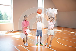 Little cute cheerleaders standing near classmate with ball