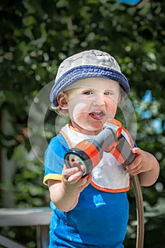 Little cute child in blue hat holding a garden hose outdoors sunny summer day.
