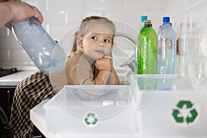 Little cute child helps mom sort plastic in the kitchen. Cute daughter puts plastic bottles in a box with a recycling