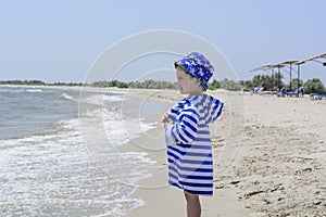 Little cute child standing on the shore in a striped dressing gown and looking at the sea, smiling, happy,