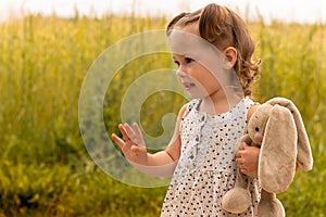 Little cute girl 1-3 with a plush hare in a light dress waving his hand in a field of spikelets of rye