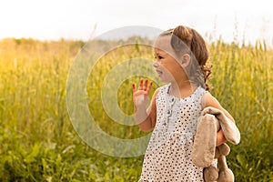 Little cute girl 1-3 with a plush hare in a light dress waving his hand in a field of spikelets of rye