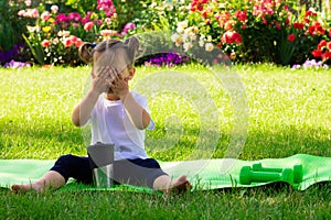 Little cute girl 1-3 in a white t-shirt drinks water after sports, sitting on a green mat on a background of flowers