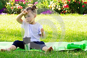 Little cute girl 1-3 in a white t-shirt drinks water after sports, sitting on a green mat on a background of flowers