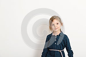 Little cute girl 4-6 years old wearing a blue dress with polka dots stands against the background of a white wall