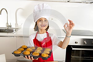 Little and cute girl alone in cook hat and apron presenting and showing tray with muffins smiling happy