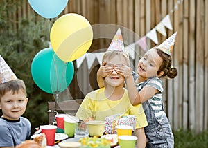 Little cute girl from behind closes eyes brother or friend with hands. Cute funny nine year old boy celebrating his birthday with