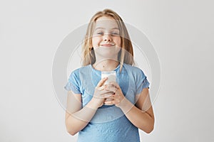Little cute girl with blonde hair in blue t-shirt with milk drops on face, happy to start her day with big glass of