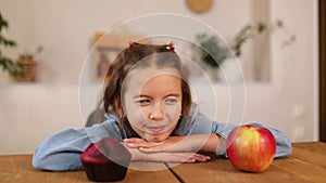 Little cute girl choosing between sweet cake and fresh red apple in kitchen at home