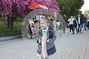 Little cute girl in a coat walks in the park against the background of a flowering tree in spring