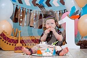 Little cute girl in costume of Apache Indian chief and in a feather headdress sits and tastes a sweet cake. First birthday 1 year