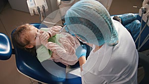 Little cute girl in the dentist`s office does not want to be treated, smiling, resisting and closing her mouth with her