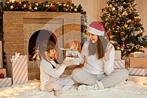 Little cute girl giving present to her mother, excited woman getting gift box, family sitting on floor on soft carpet near