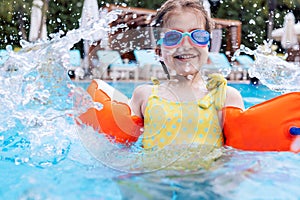 Little cute girl in goggles, yellow swimsuit and orange inflatable armlets swiming in pool hotel