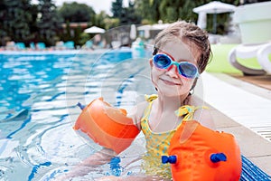 Little cute girl in goggles, yellow swimsuit and orange inflatable armlets swiming in pool hotel